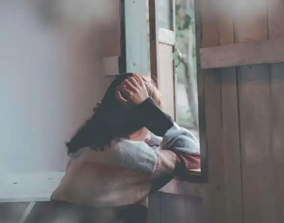 Person sitting by an open window with head in hands, expressing sadness or deep thought in a quiet room